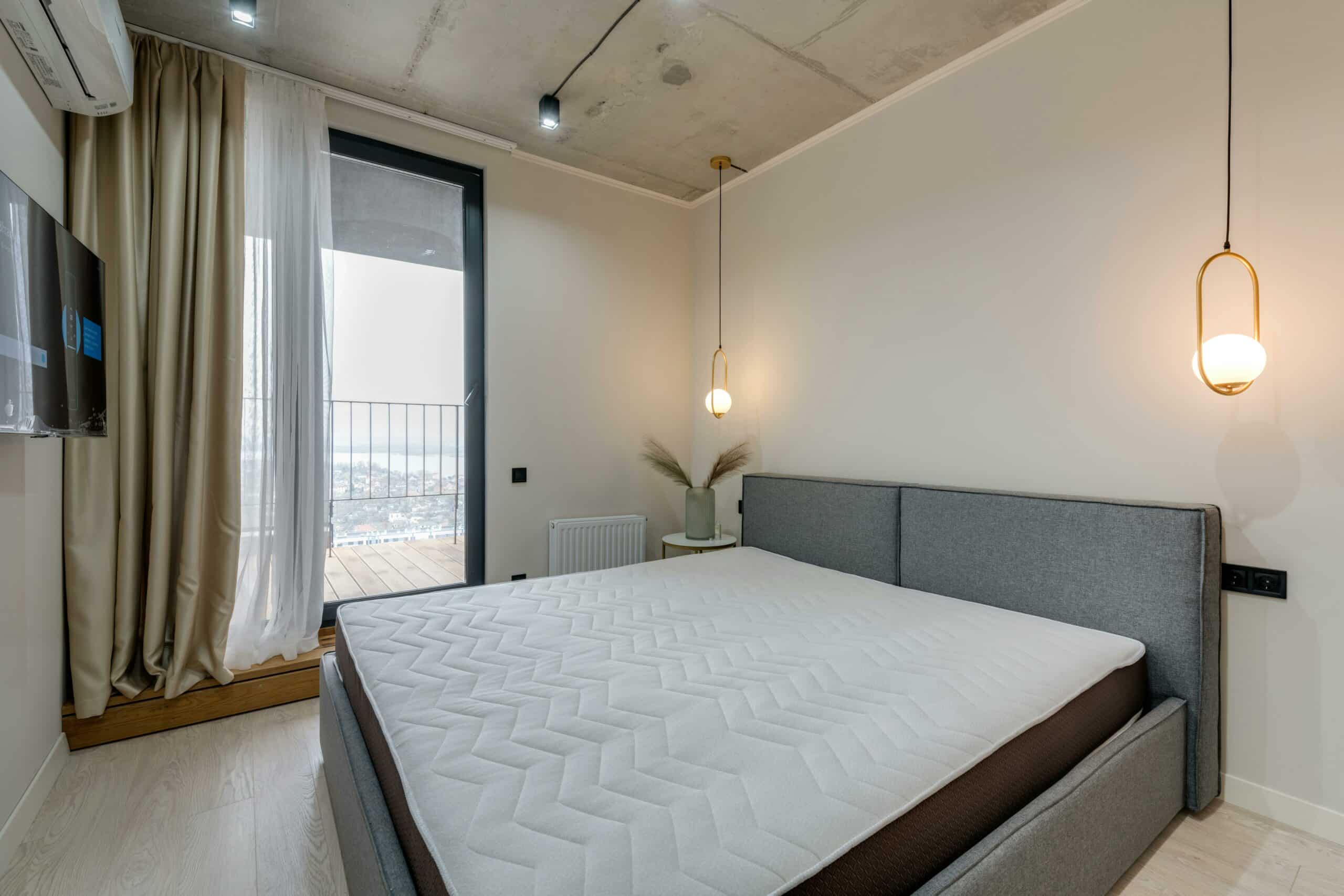Vue d'une chambre d'hôtel moderne avec un grand lit au cadre en tissu gris et un matelas épais au matelassage géométrique blanc. Deux suspensions dorées encadrent le lit devant un mur uni et une baie vitrée.