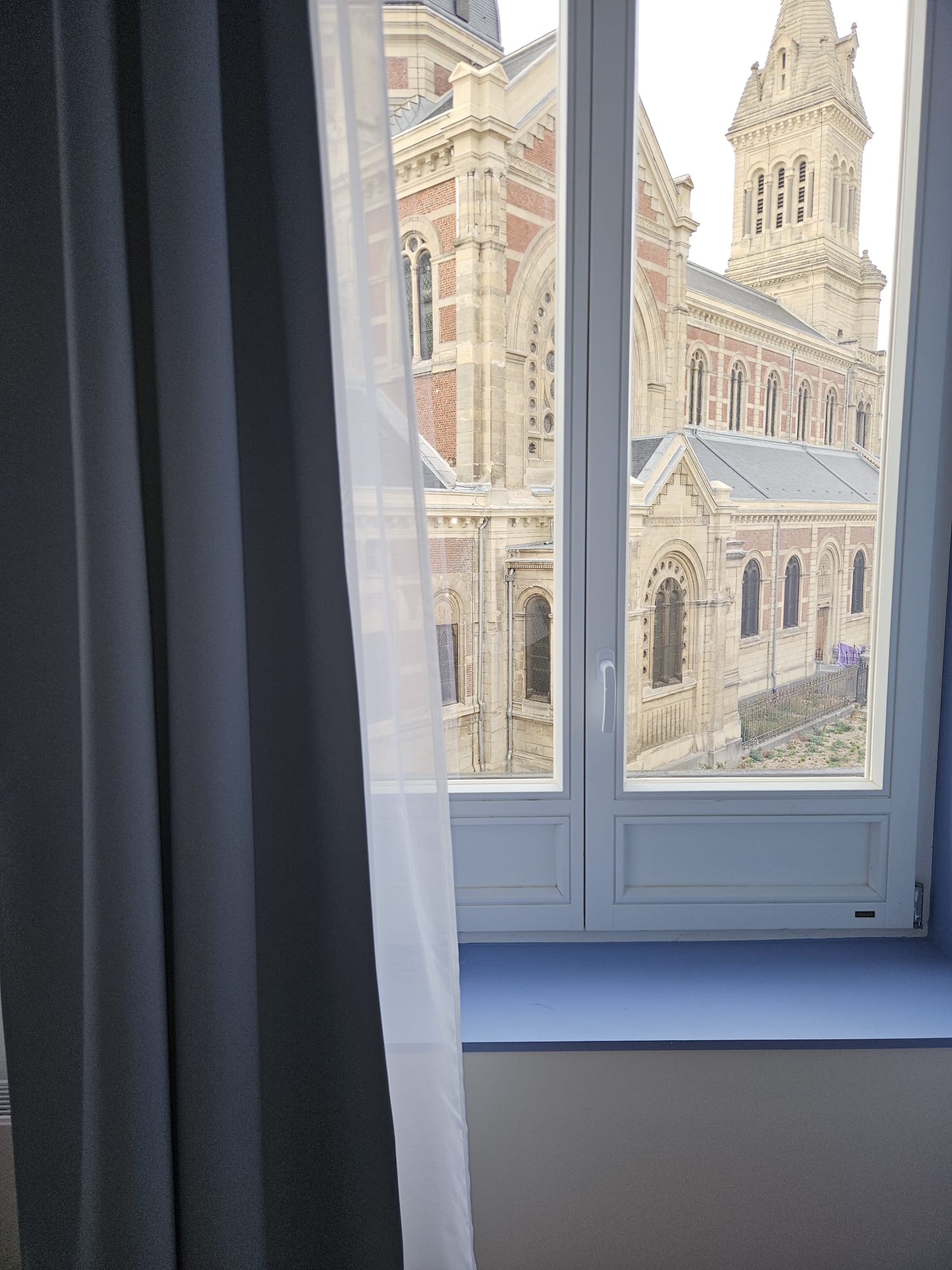 Voilage blanc et rideau bleu encadrant une fenêtre avec vue sur un bâtiment historique à Lille