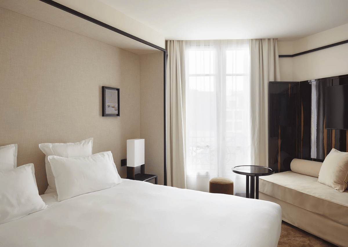 Chambre de l’Hôtel Atlas à Paris avec lit blanc, voilage blanc, rideaux beiges et espace salon.